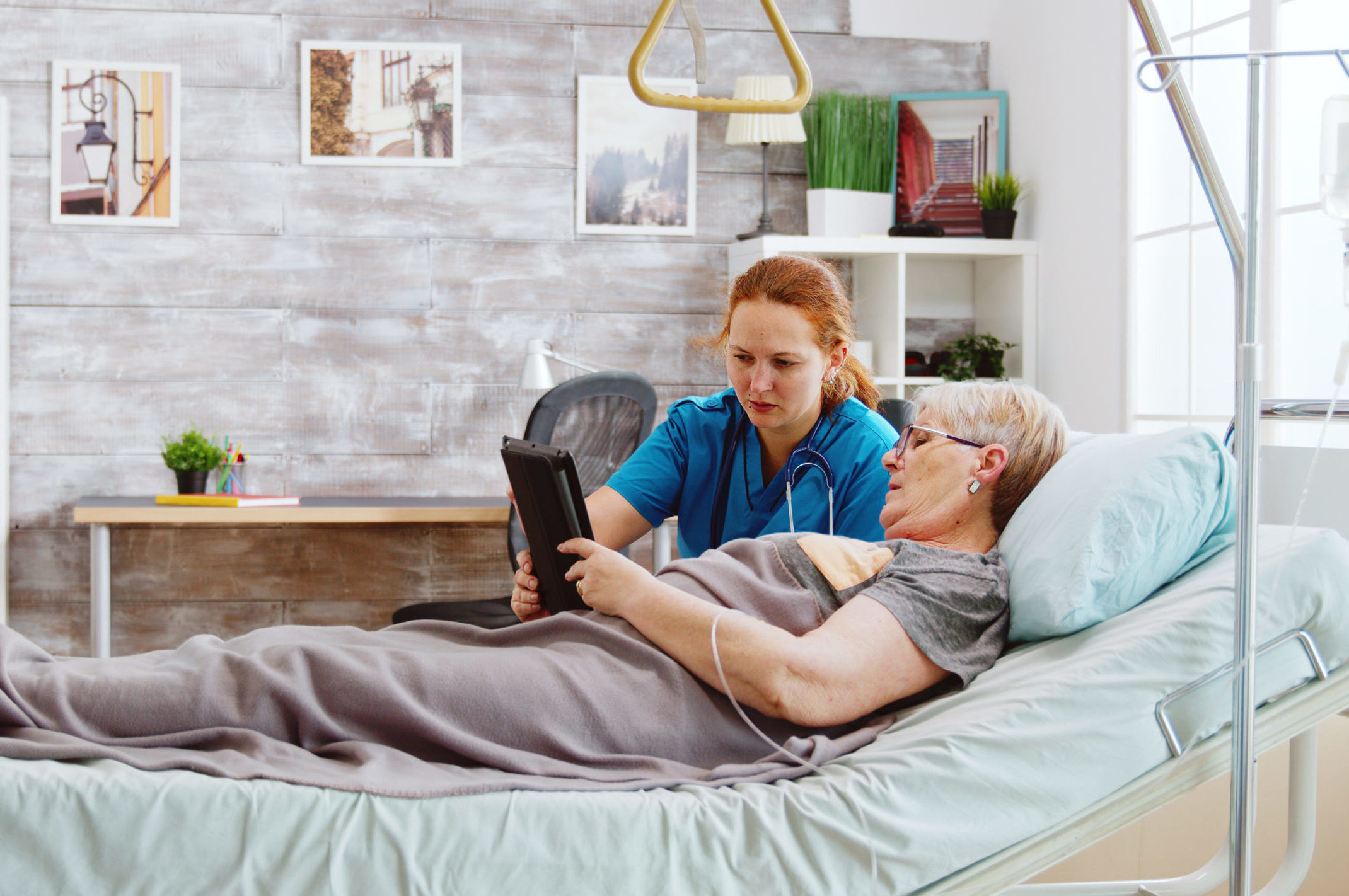 Female caregiver helps an old disabled woman lying in hospital bed to use a digital tablet PC. Bright room with big windows