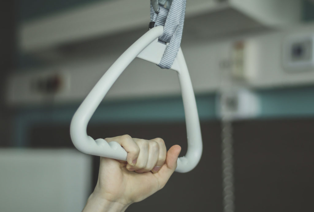 Caucasian male patient holding with one hand on a triangular therapeutic arch with a gripper set above the bed in a hospital room, close-up side view. The concept of trauma devices.