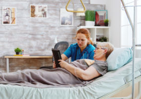 Female caregiver helps an old disabled woman lying in hospital bed to use a digital tablet PC. Bright room with big windows