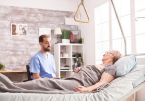 Male doctor talking with retired senior woman in nursing home lying in bed.